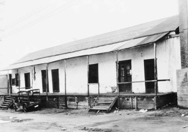 Photograph of an exterior view of the Avila Adobe Homestead on Olvera Street, June 25, 1928. The long, wooden structure stands at center with the corner of a second, brick structure to the right of it. The adobe has a wooden porch which stands several feet above the dry, dirt ground. Two small staircases reach from the ground to the porch where several lightly-colored doors and windows stand in the dark walls. A small automobile is parked in front of the adobe on the left while electrical lines extend above the adobe's roof.