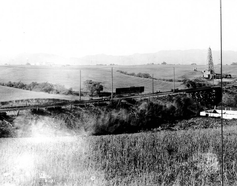 Photograph of a panoramic view of  farmland on Santa Monica Boulevard looking east, 1904. At left, a farmhouse which includes a windmill and corral can be seen standing to the right of a long road which continues into the distance. Closer to center, the roof of another building on the homestead can be seen behind a foregroudn hill. The road is lined by utility poles. Oil wells speckle the distance, where a mountain can be seen faintly to the left.