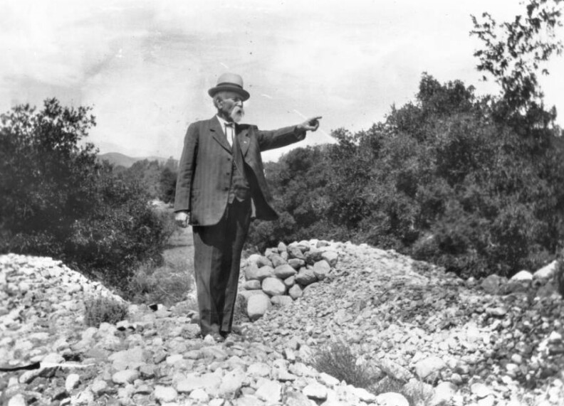 Prudhomme, who is wearing a striped jacked, vest, shirt, trousers, and bowler hat, points to his left. He has a large white mustache and goatee. He is standing among small piles of rocks. Bushes grow behind him.