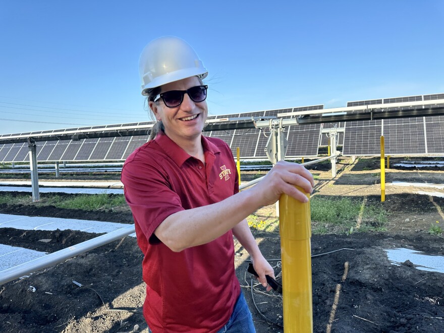 A man in a hard hat and red shirt stands in front of solar panels. Matt O’Neal, an entomology professor at Iowa State University, shows off the school’s experimental solar farm just south of Ames.